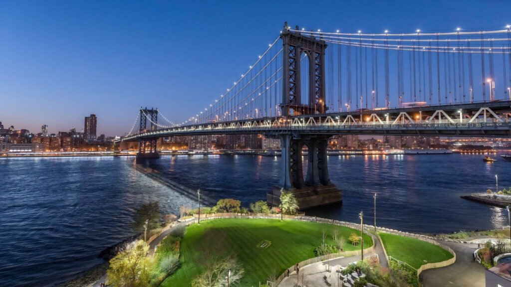 Couples relaxing on the waterfront lawn at Brooklyn Bridge Park with Lower Manhattan skyline at sunset.